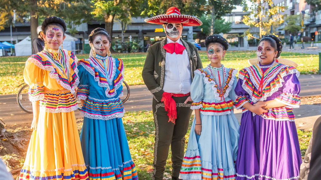 Four women dressed in traditional Mexican La China Poblanca dresses and a man dressed in a traditional Charro outfit. They all have face paintings representing La Catrina.