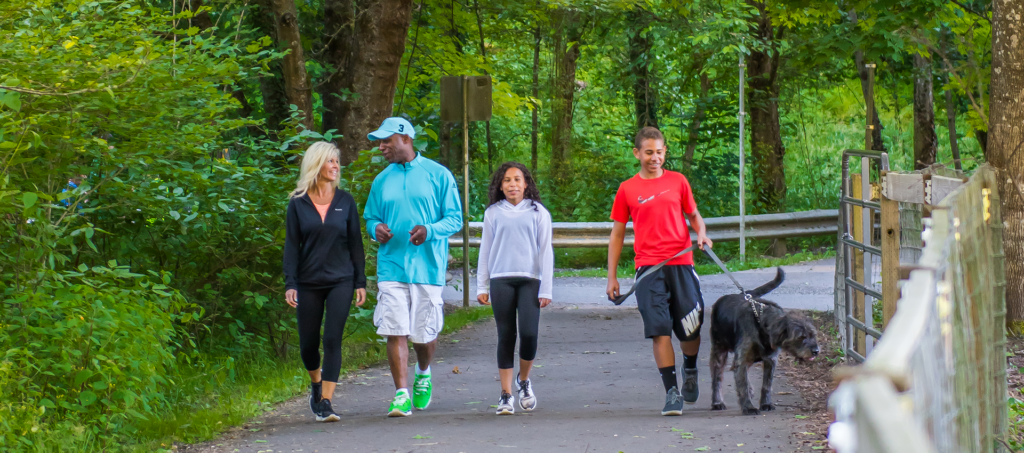 A family of four with two parents and two children walking their dog on a paved trail.