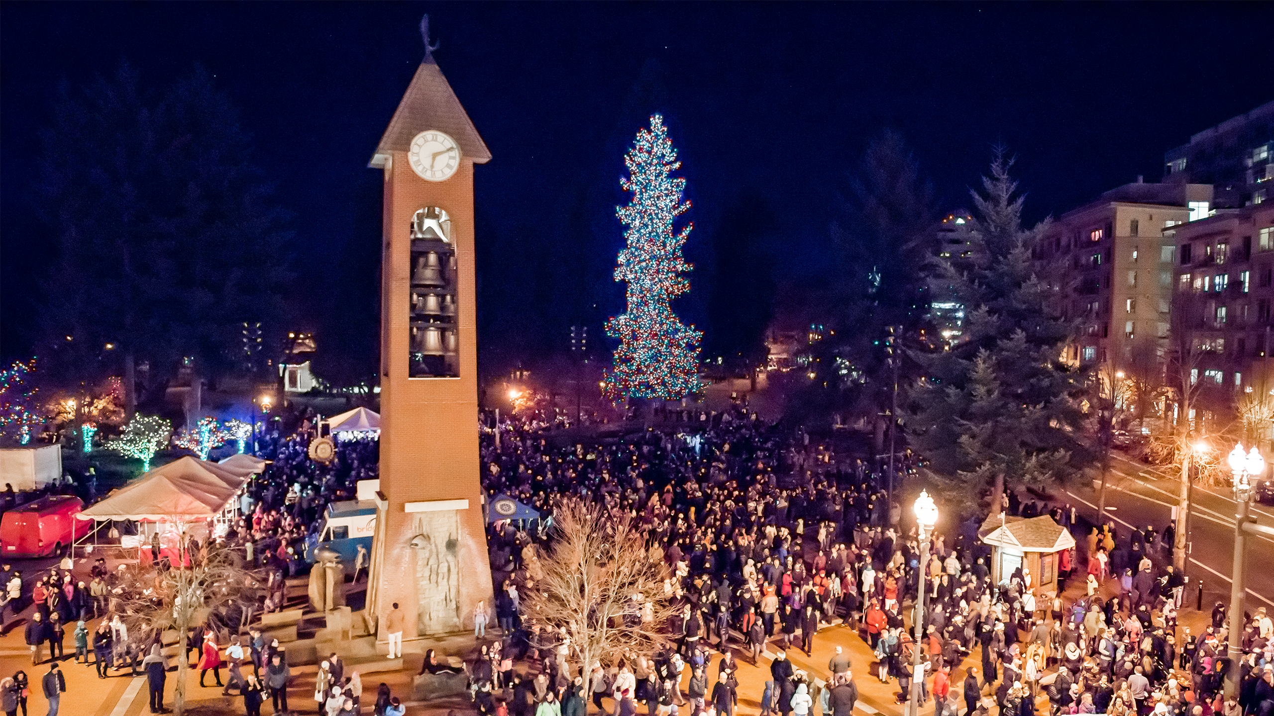 An aerial view of Propstra Square at night. It is filled with people, the clock tower in the foreground and a lit holiday tree in the background.