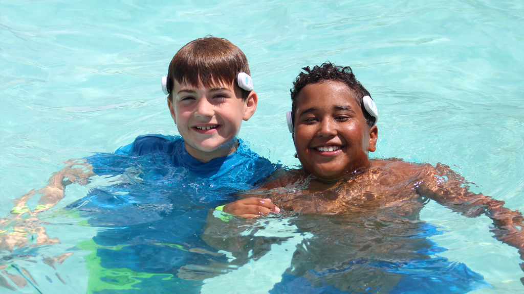 Two children in a pool smiling at the camera while wearing WAVE drowning prevention headsets.