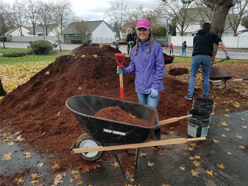 A volunteer in a purple jacket poses with a shovel next to a mulch pile and wheelbarrow