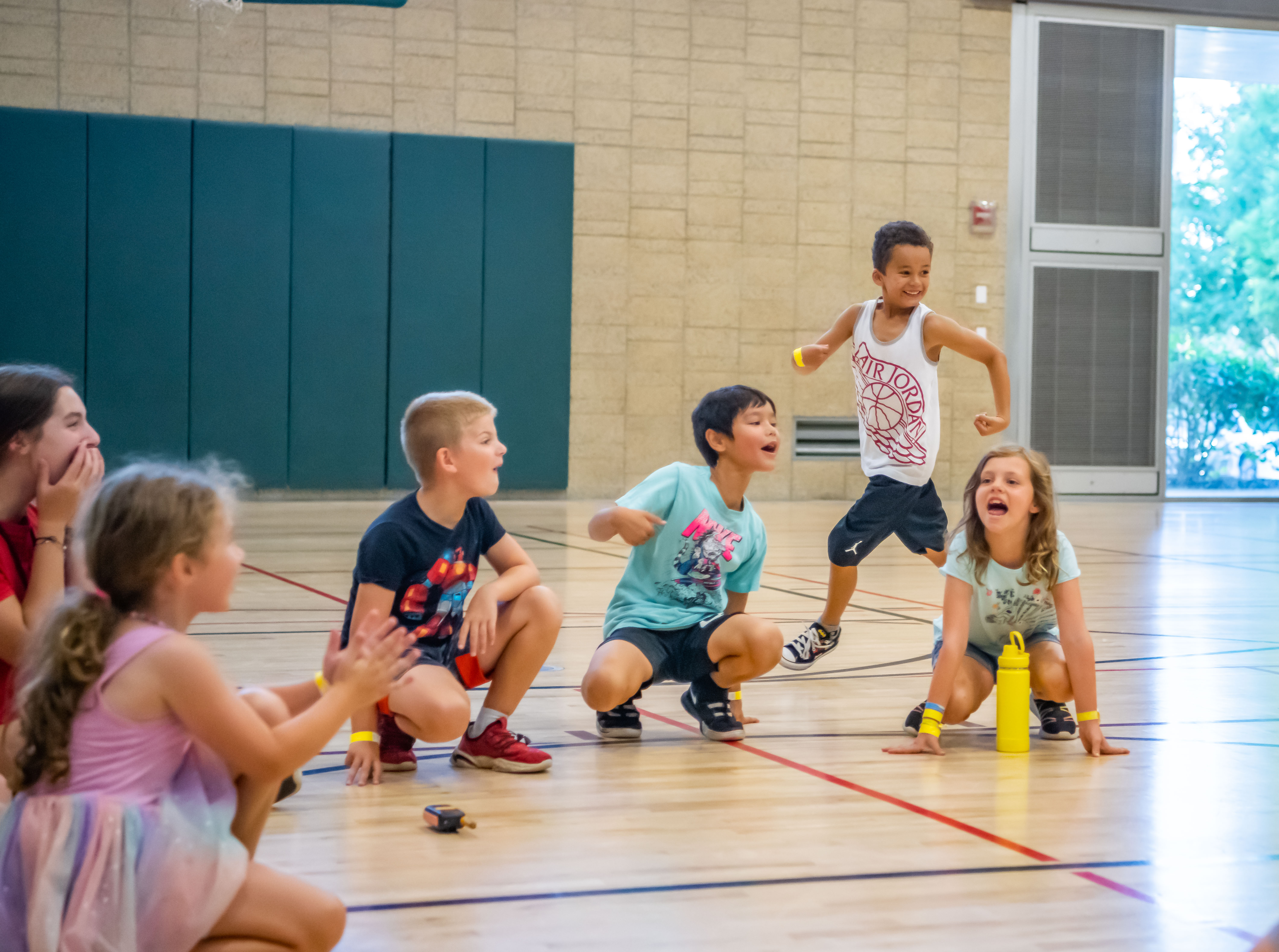 A group of young kids sitting on a gym floor, playing duck-duck-goose