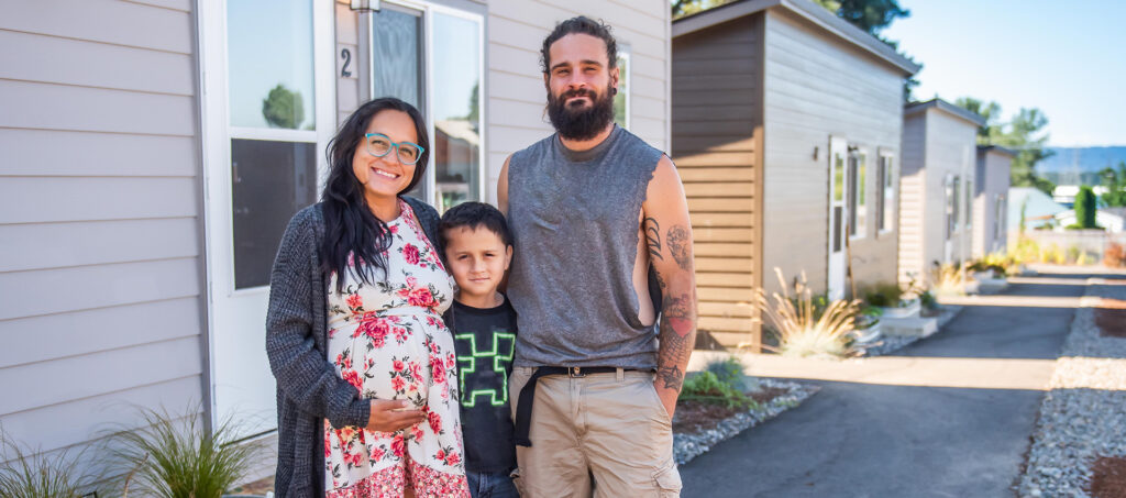 Two parents, one child standing in front of their tiny home, the community was built thanks in part to the Affordable Housing Fund