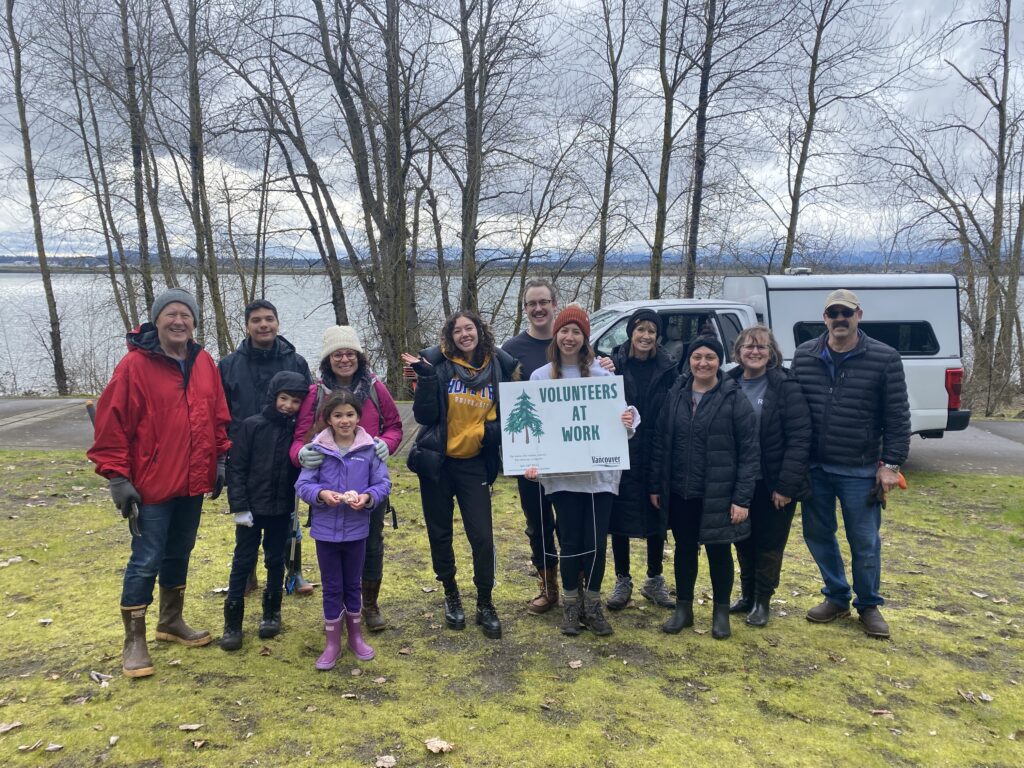 group of adult and child volunteers pose on the grass with the Columbia River in the background and trees in winter