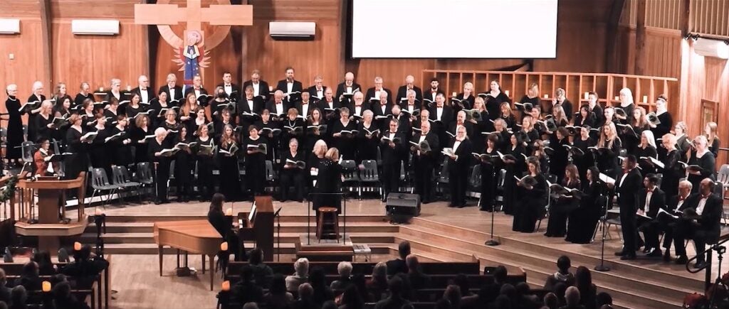 A large choir dressed in black sings in a church, accompanied by an orchestra. 