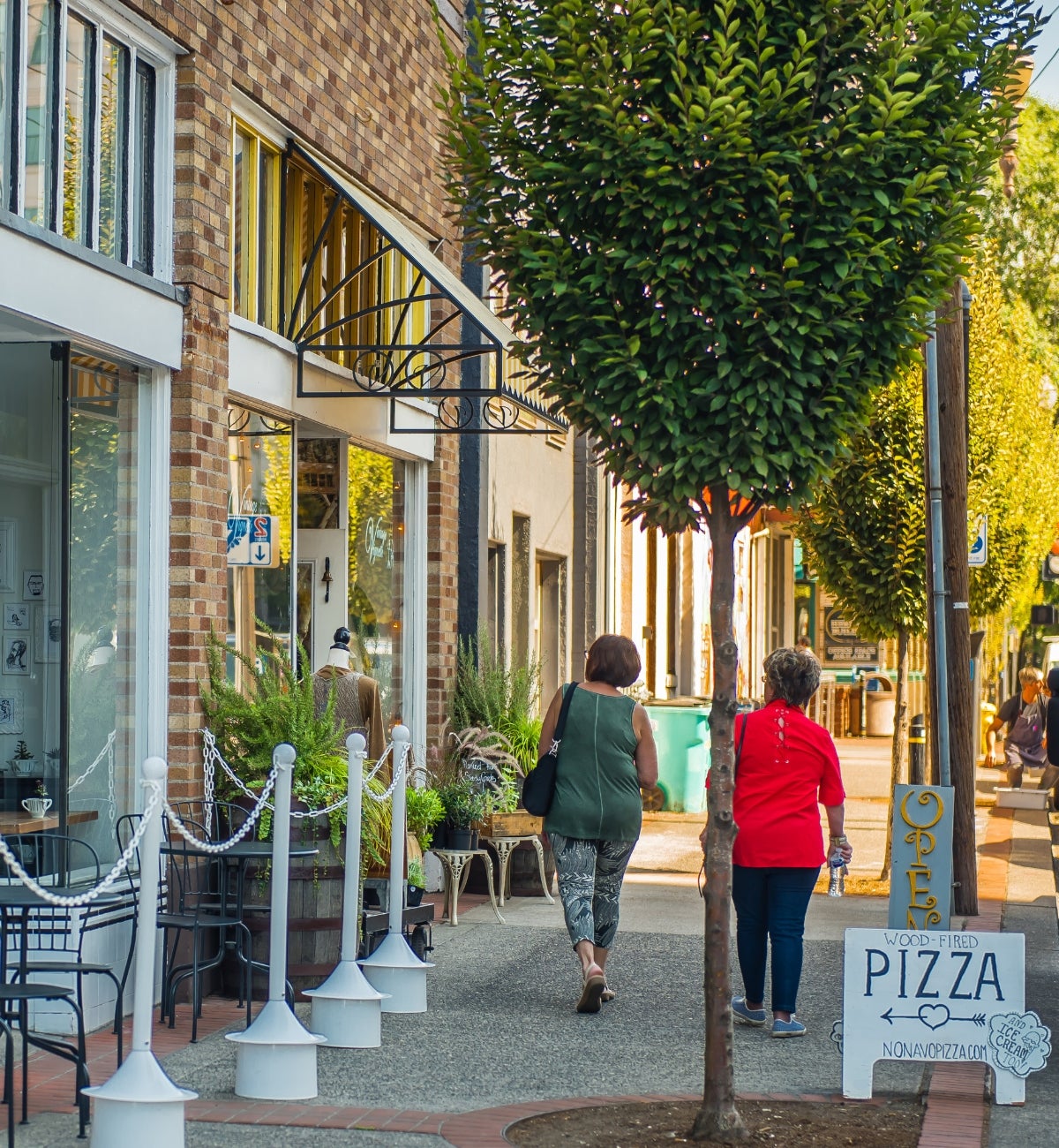 women walking downtown in front of small businesss store fronts and a pizza sidewalk sign