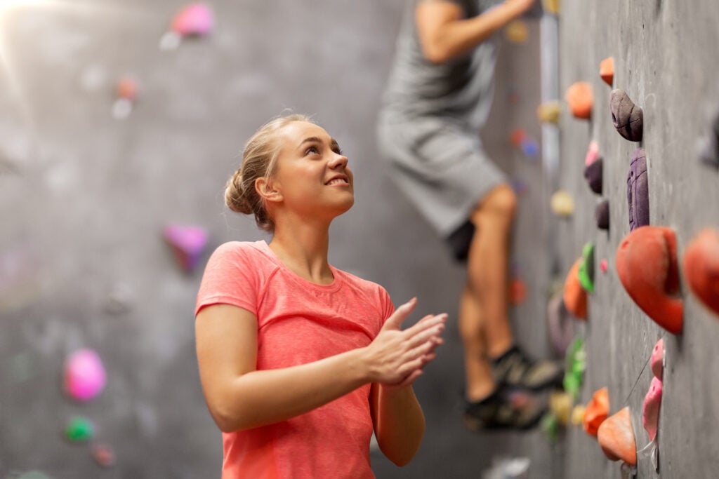 A young woman smiles and looks up at a rock climbing wall, another person is already climbing the wall behind her.