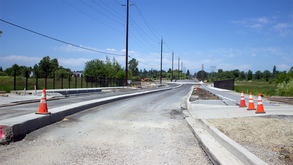 Active construction site along NE 137th Ave, with concrete installed for center lane divider, gravel and pavement along new road, orange cones in middle and to the side of the street