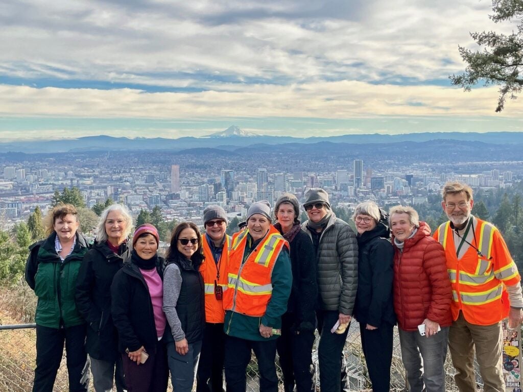 A group of smiling older adults on the Pittock Mansion hike with Portland and Mt. Hood behind them.