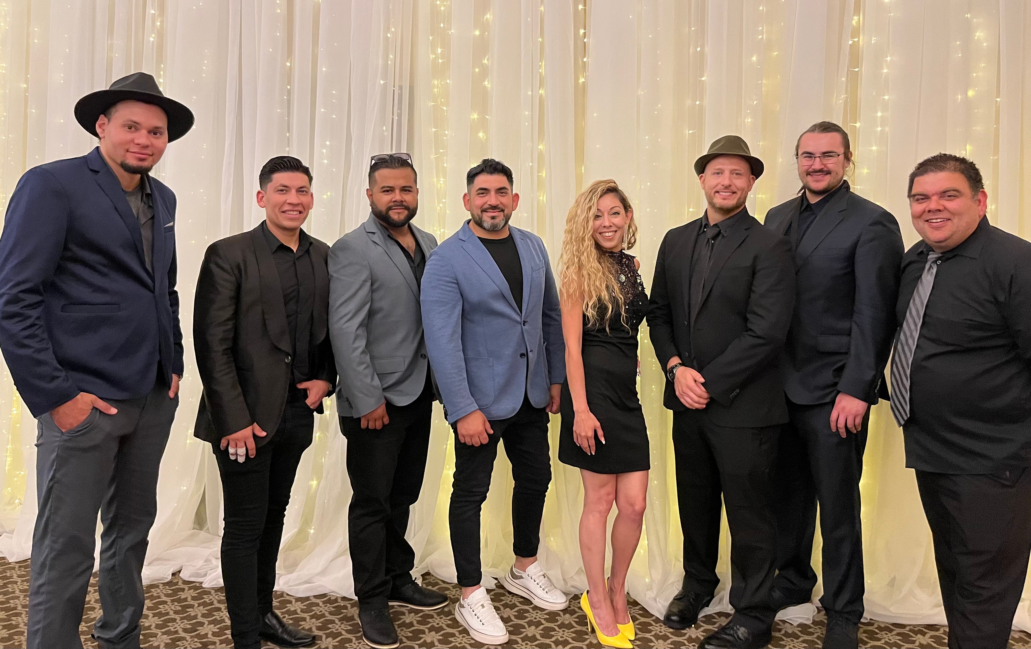 The Cuban band, Dina y los Rumberos, pose together in front of a string light backdrop.