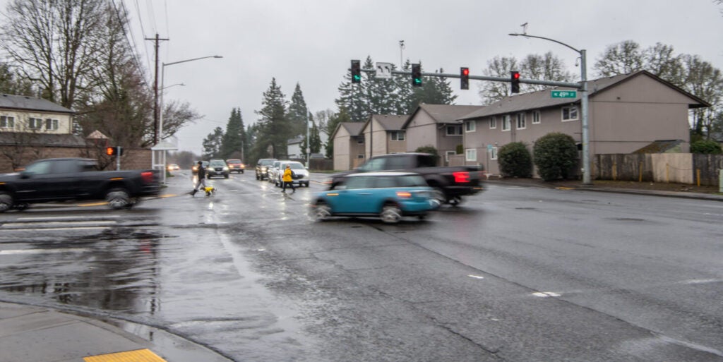112th Avenue at 49th Street. Pedestrians cross 112th Street. Vehicles are turning off of 112th street onto 49th street. It's raining out
