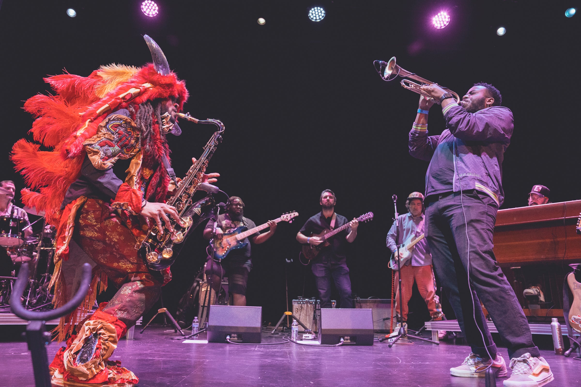 The band Cha Wa plays brass instruments on stage while dressed in colorful traditional Mardi Gras Indian outfits.