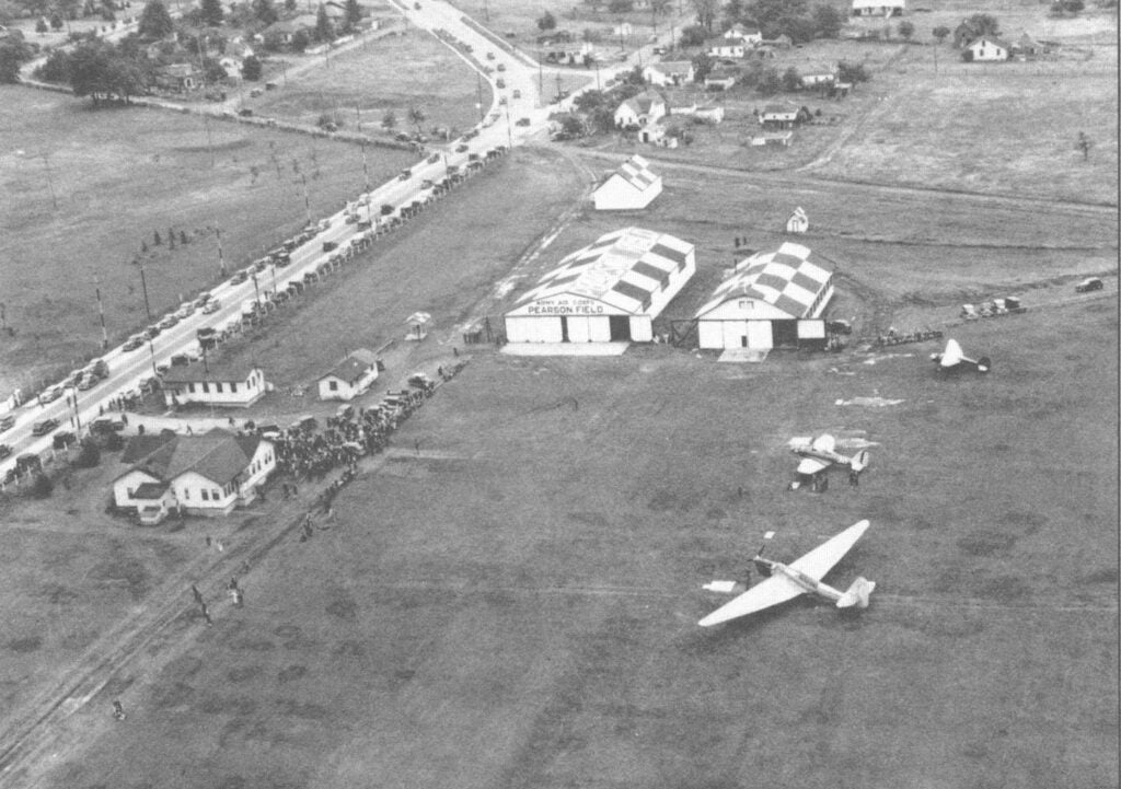 Historic overhead of Pearson Field, with a crowd of people near a building and three planes in the field.