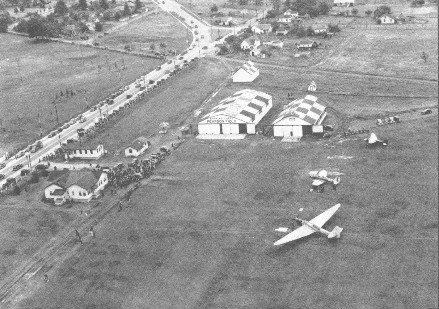 Historic overhead of Pearson Field, with a crowd of people near a building and three planes in the field.