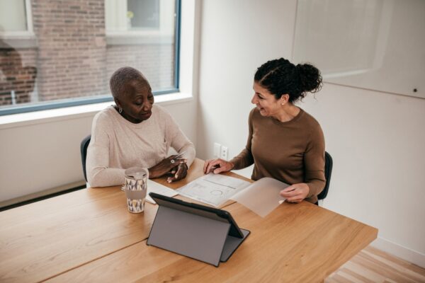 Two women sit together at a table with a tablet and paperwork.