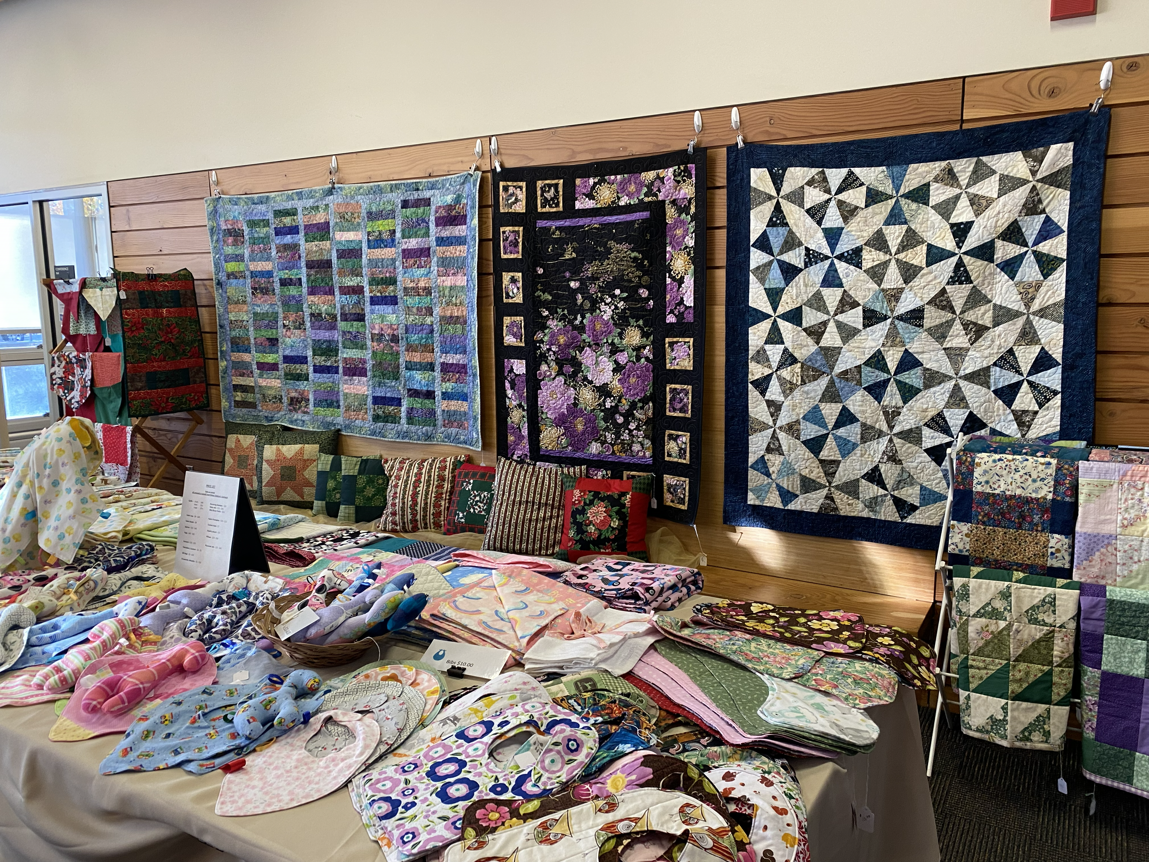 A table full of colorful handmade bibs and baby blankets stands in front of a wall displaying a variety of quilts.