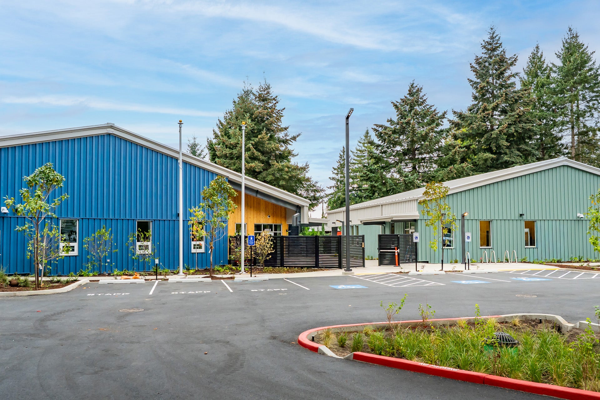The front of the bridge shelter. One blue building on the left and a teal building on the right. ADA and staff parking line the front of the gated campus.